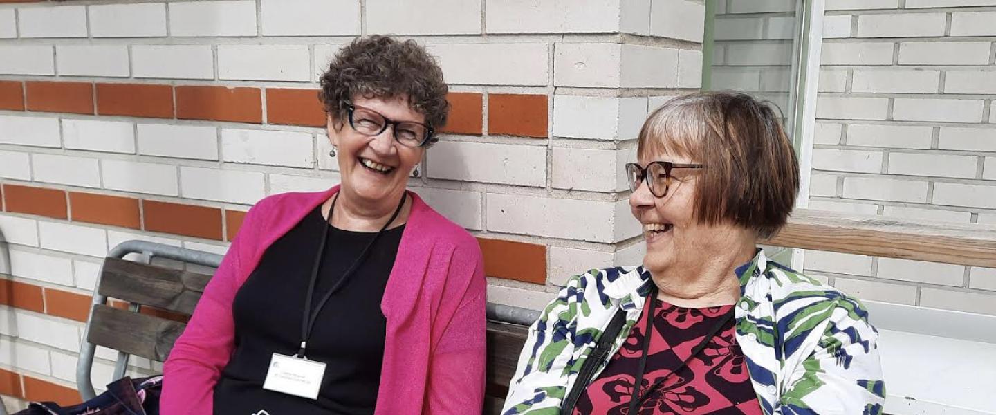 Kaksi penkillä istuvaa iäkästä nauravaa naista katsovat toisiinsa